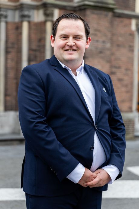 Een man in een donkerblauwe blazer en een wit overhemd staat met gekruiste armen lachend buiten voor een gebouw.