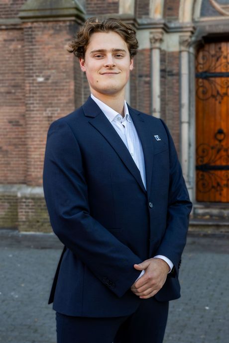 Een man in een donkerblauwe blazer en een wit overhemd staat met gekruiste armen lachend buiten voor een gebouw.