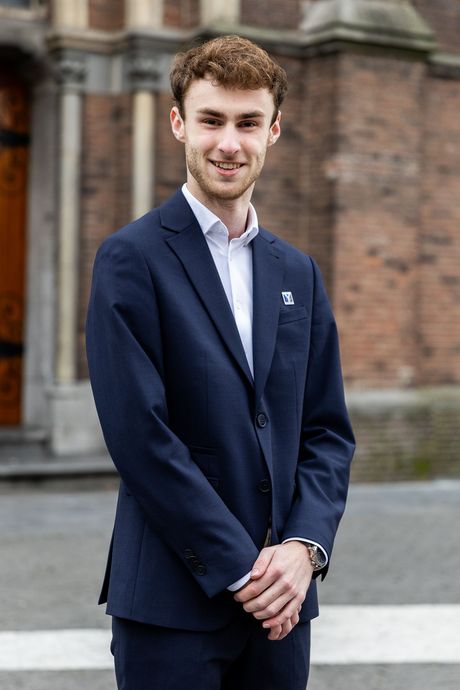 Man in marineblauw pak, wit overhemd, glimlachend, handen ineengevouwen, buiten voor een bakstenen gebouw.