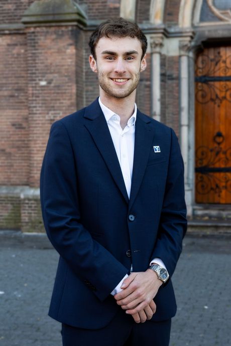 Een jonge man in een donkerblauw pak glimlacht, met zijn handen ineengevouwen, voor een bakstenen gebouw.
