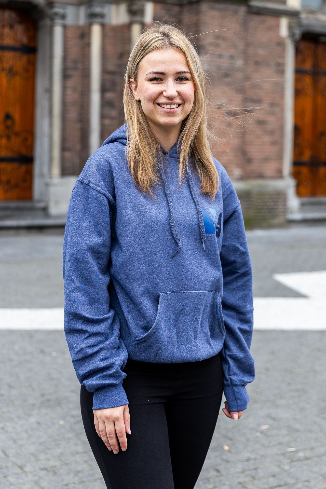 Vrouw met blond haar, gekleed in een blauwe hoodie en zwarte broek, lacht buiten.