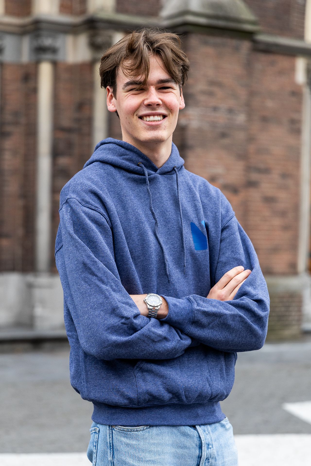 Jongere in blauwe hoodie, armen over elkaar, lachend, buiten voor een gebouw.