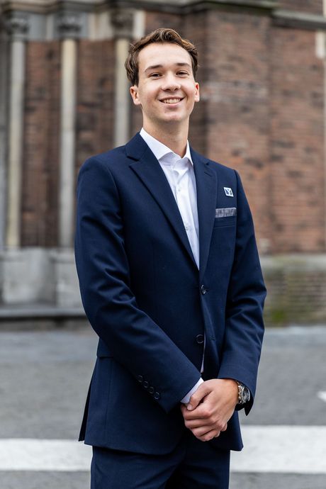 Een man in een donkerblauw pak en wit overhemd glimlacht terwijl hij voor een bakstenen gebouw staat.