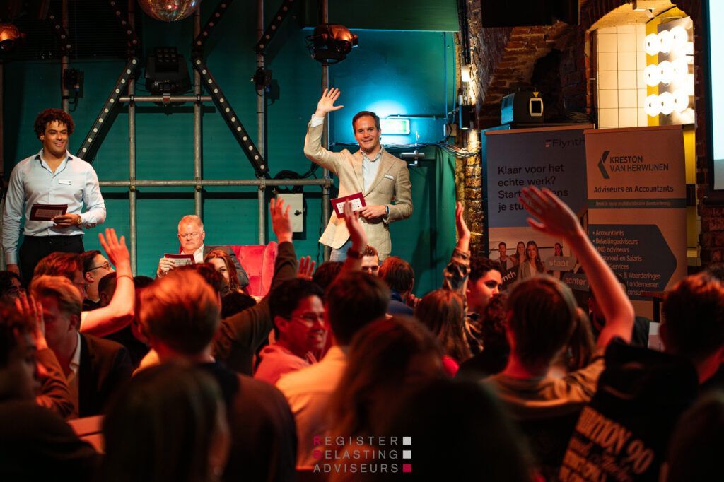 Een menigte bij een levendig evenement binnenshuis kijkt naar twee sprekers op een podium, waarvan er één een arm opheft.