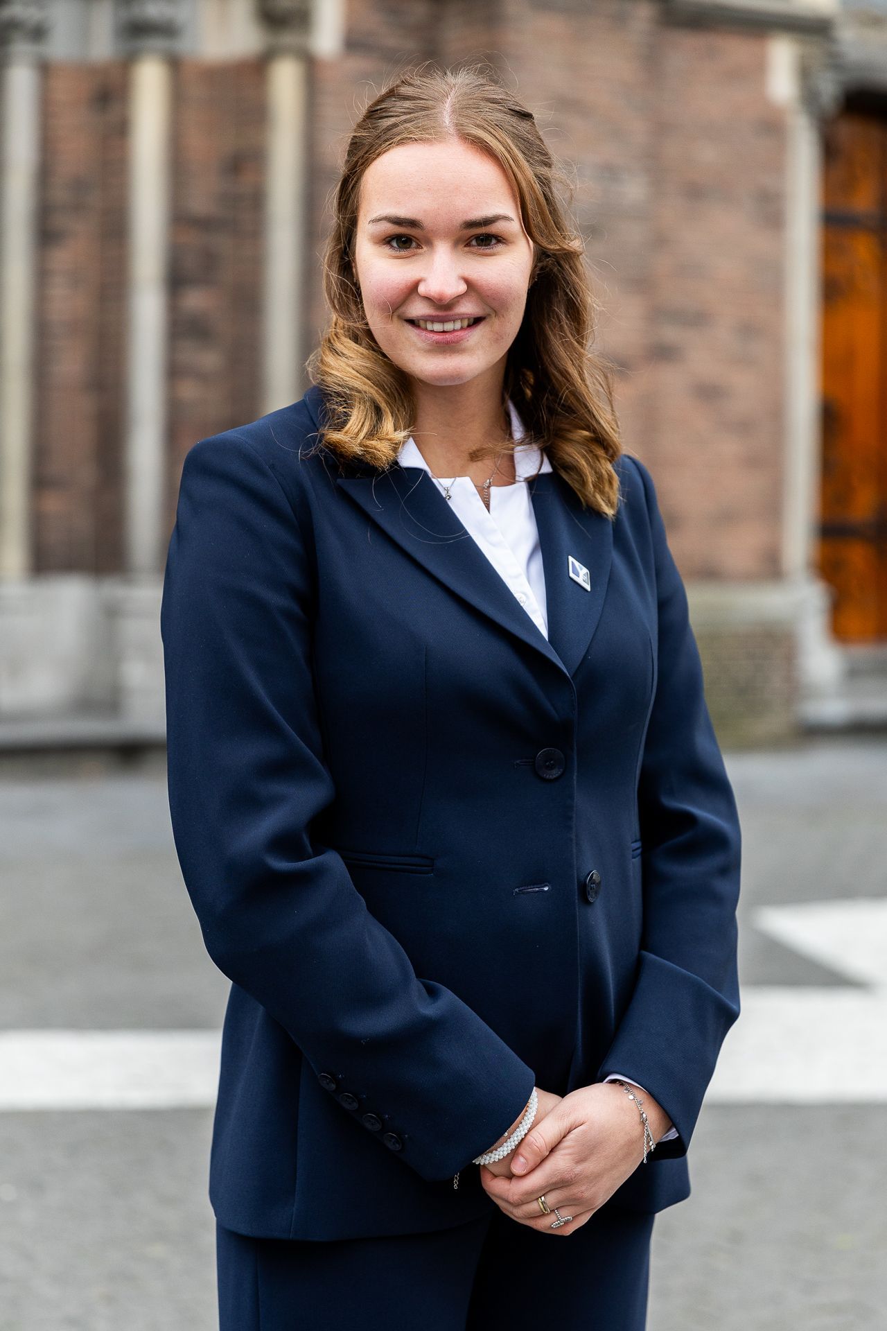 Een vrouw in een donkerblauw pak glimlacht, met haar handen ineengevouwen, buiten bij een gebouw met een houten deur.