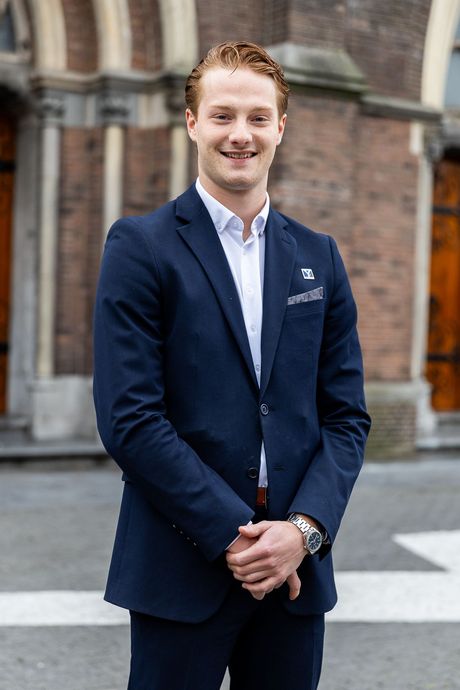 Een jonge man in een donkerblauw pak glimlacht, met zijn handen ineengevouwen, voor een bakstenen gebouw.