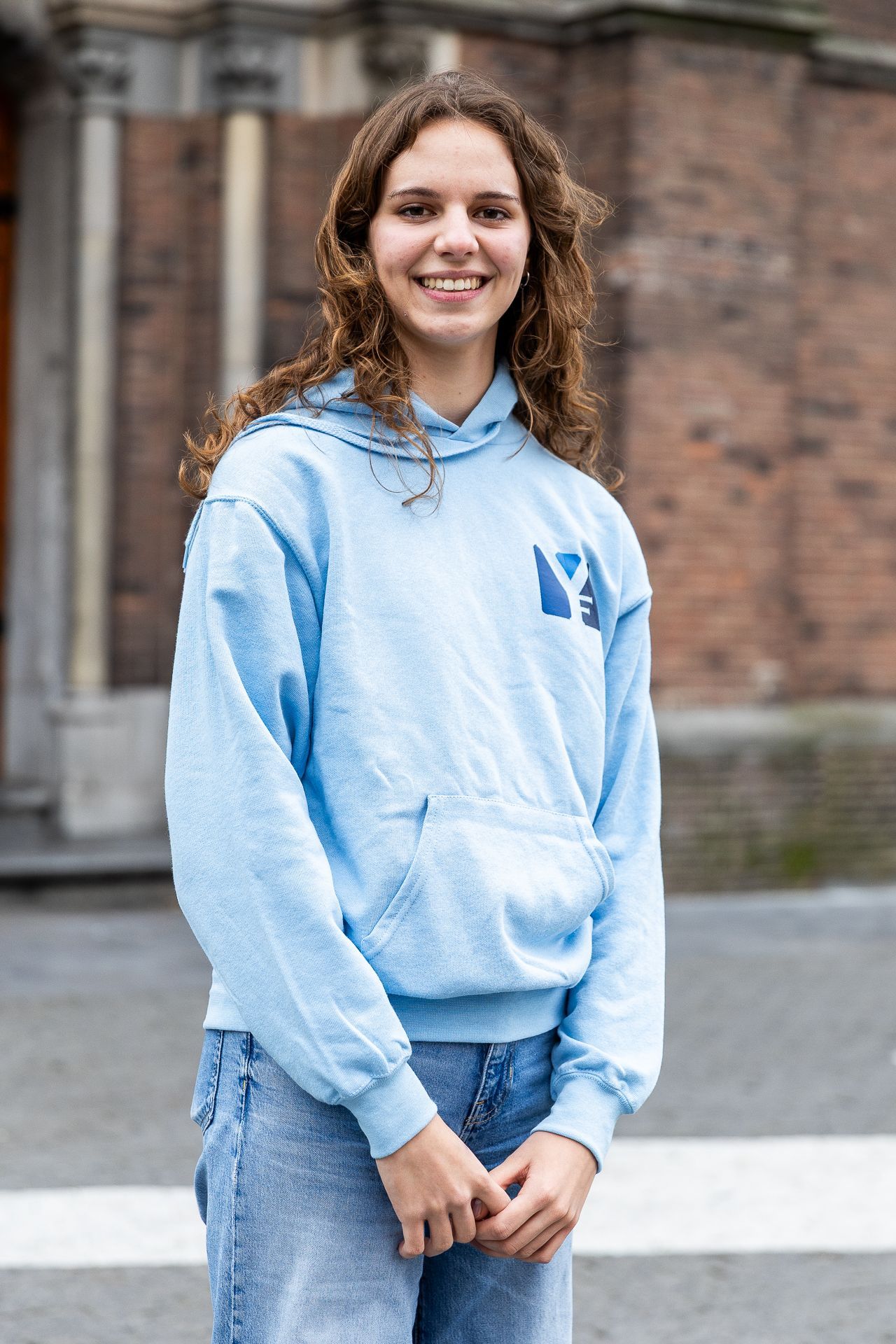 Een jong persoon in een lichtblauwe hoodie staat lachend voor een bakstenen gebouw.