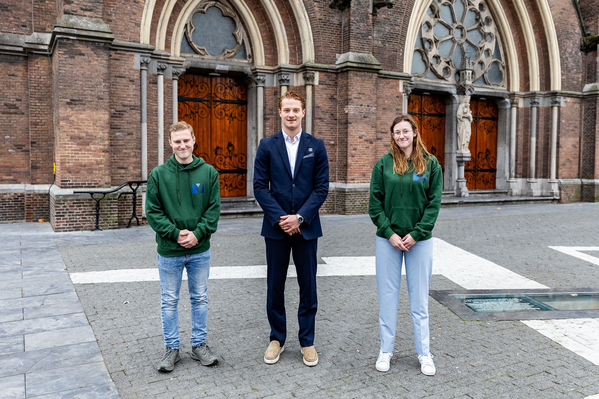 Drie mensen staan buiten een bakstenen gebouw. Twee dragen groene hoodies; één een blazer.