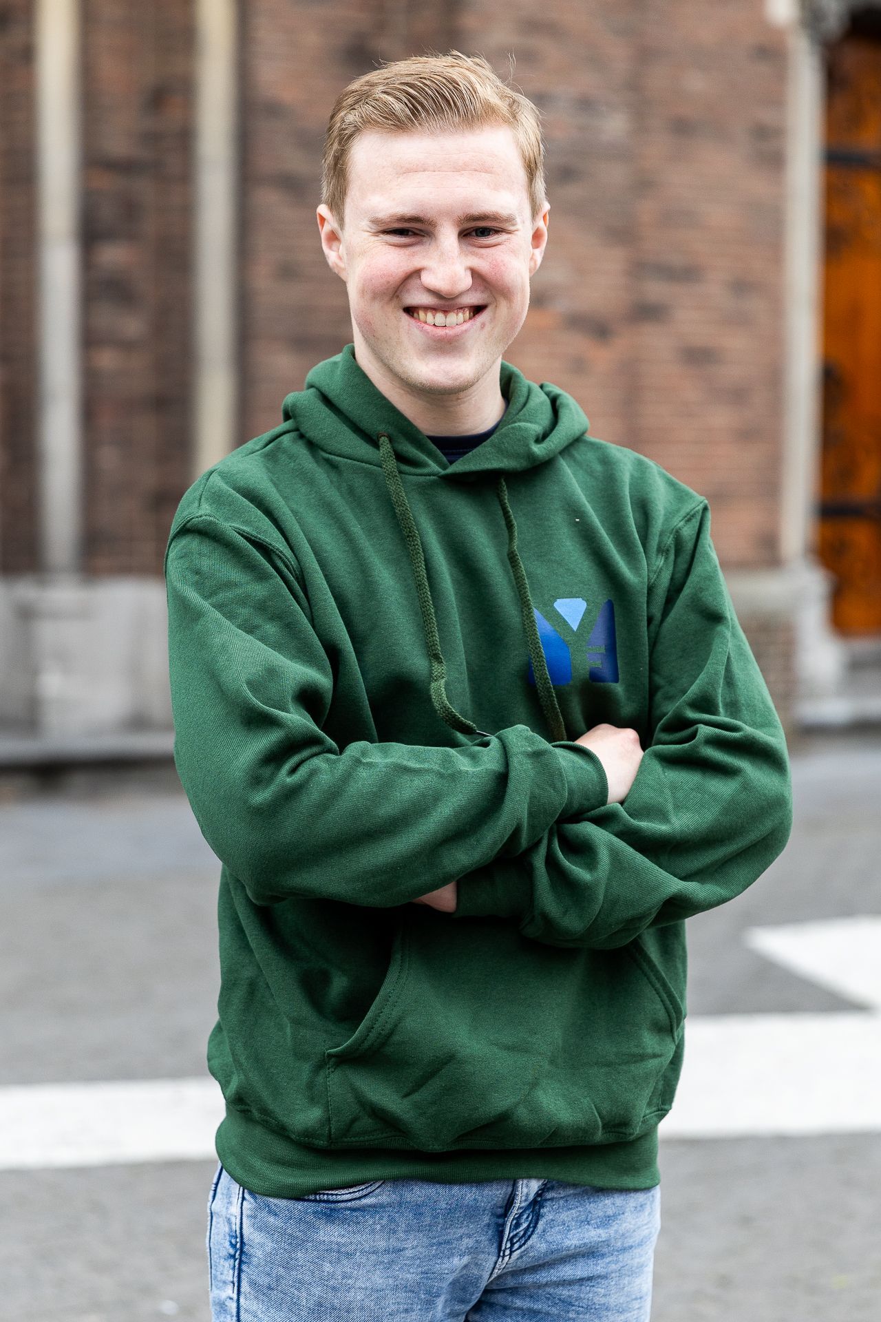 Een glimlachend persoon in een groene hoodie met de armen over elkaar staat voor een bakstenen gebouw.