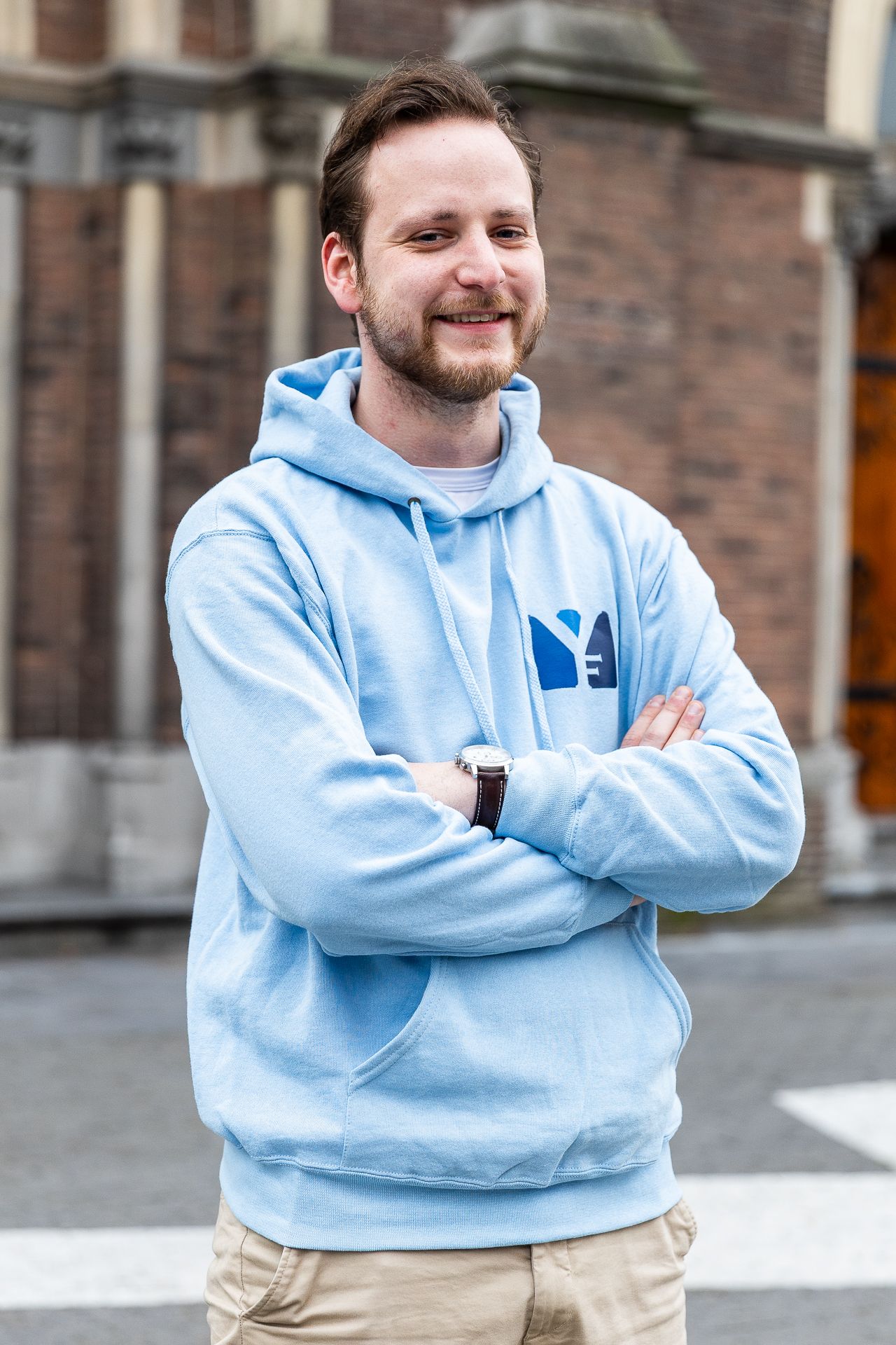 Man in lichtblauwe hoodie met gekruiste armen, lachend. Buitenomgeving.