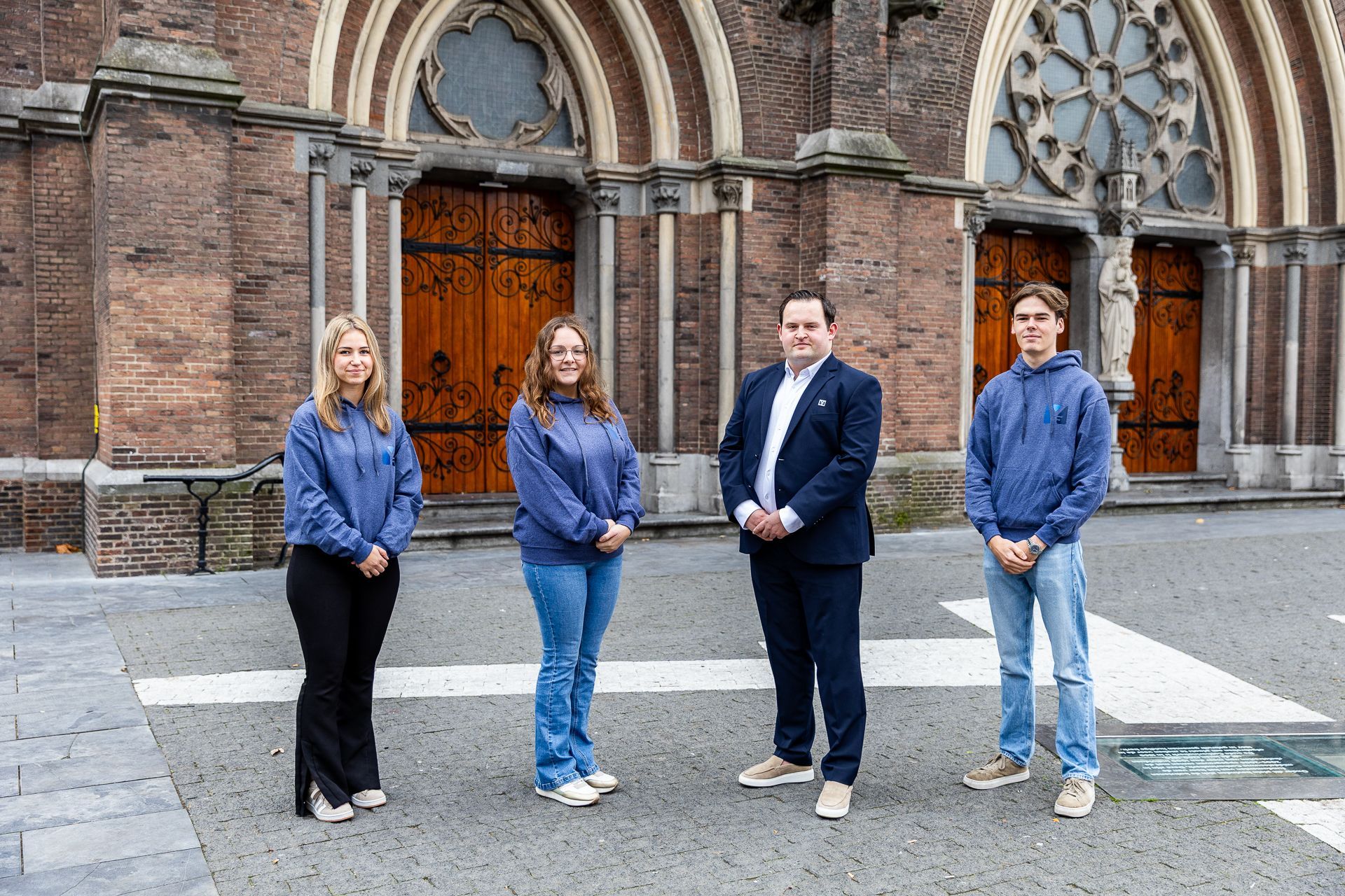 Vier mensen staan voor een bakstenen gebouw met boogvormige deuren; ze dragen allemaal blauwe shirts.