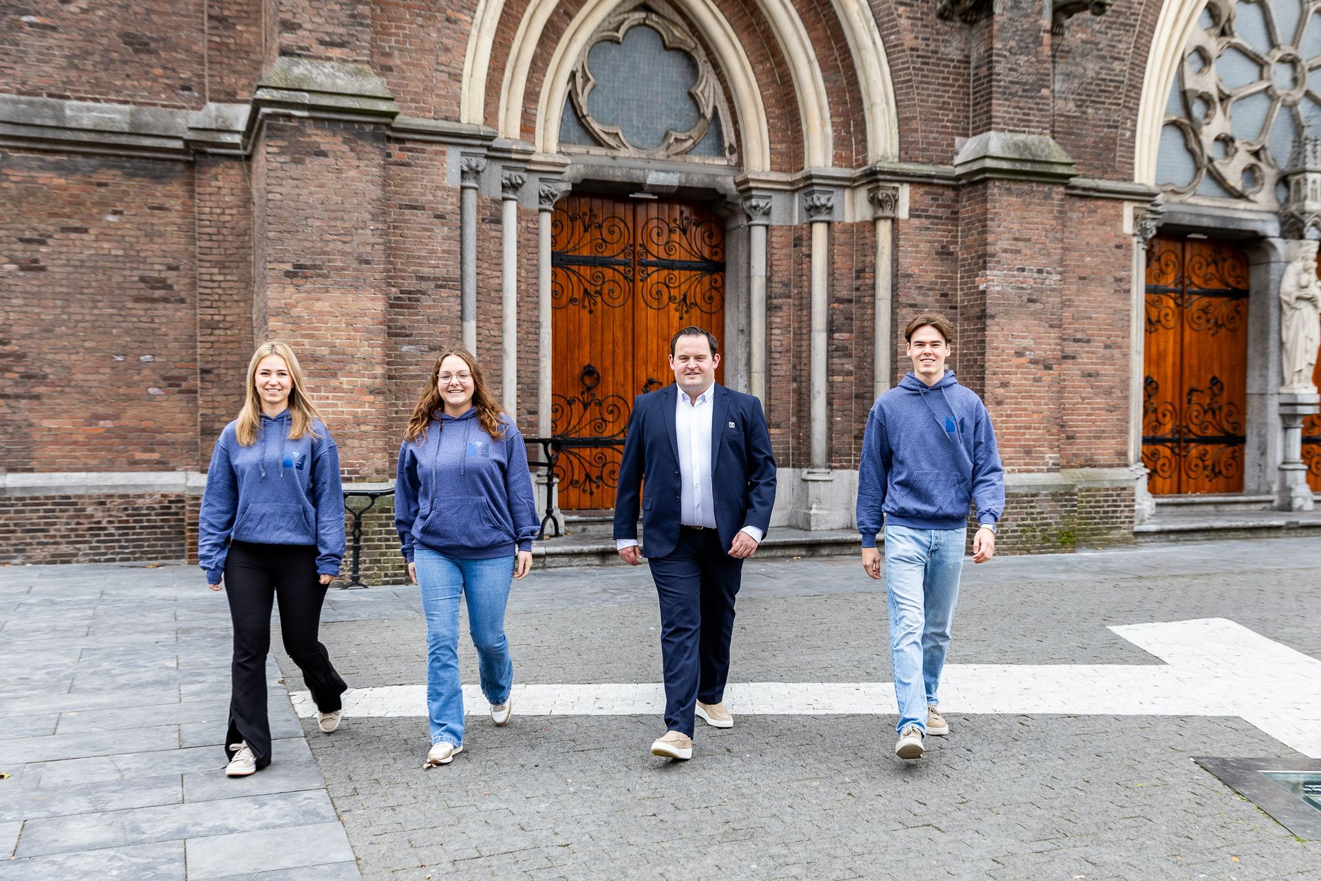 Vier mensen voor een kerk, twee met een blauwe trui aan, één in een pak, lopen in de richting van de kijker.