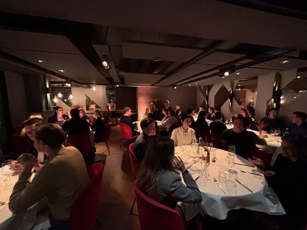 Mensen zitten aan ronde tafels in een schemerig verlicht restaurant, pratend en etend. Rode stoelen, witte tafelkleden.