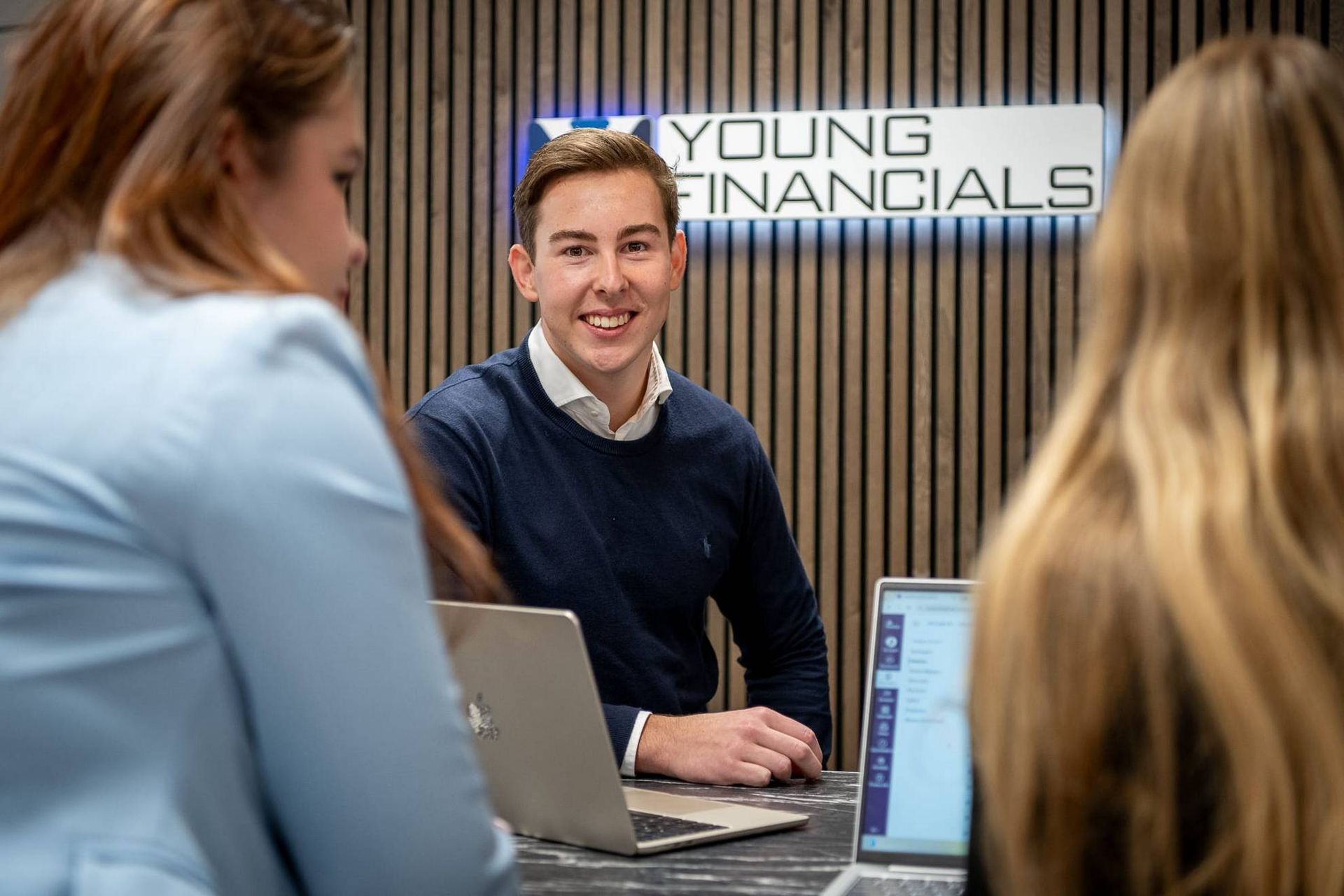 Jonge man aan een bureau, glimlachend, geflankeerd door twee vrouwen met laptops, bordje