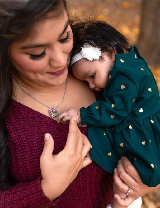 Woman in burgundy sweater holds a baby wearing a green dress with gold hearts; both are in a natural setting, looking down.