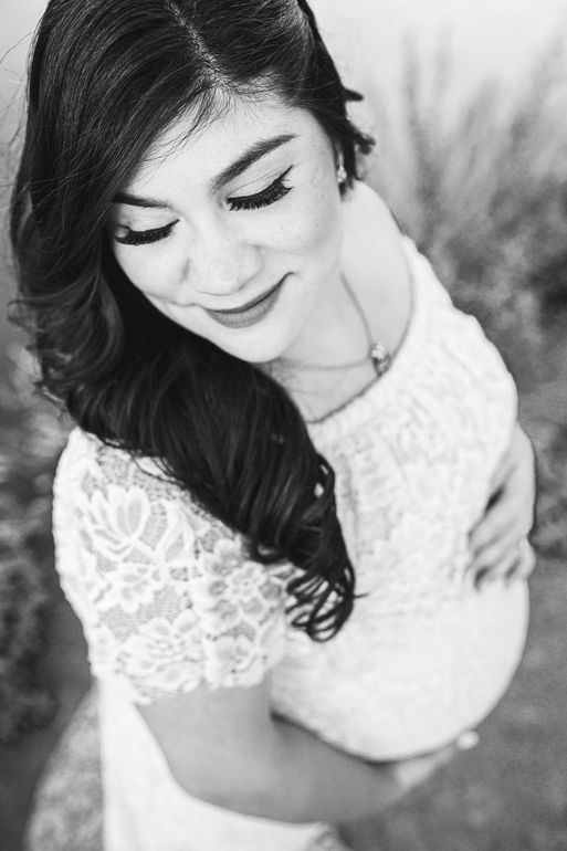 Pregnant person in a white lace dress cradles her belly, looking down with a gentle smile. Black and white photo.