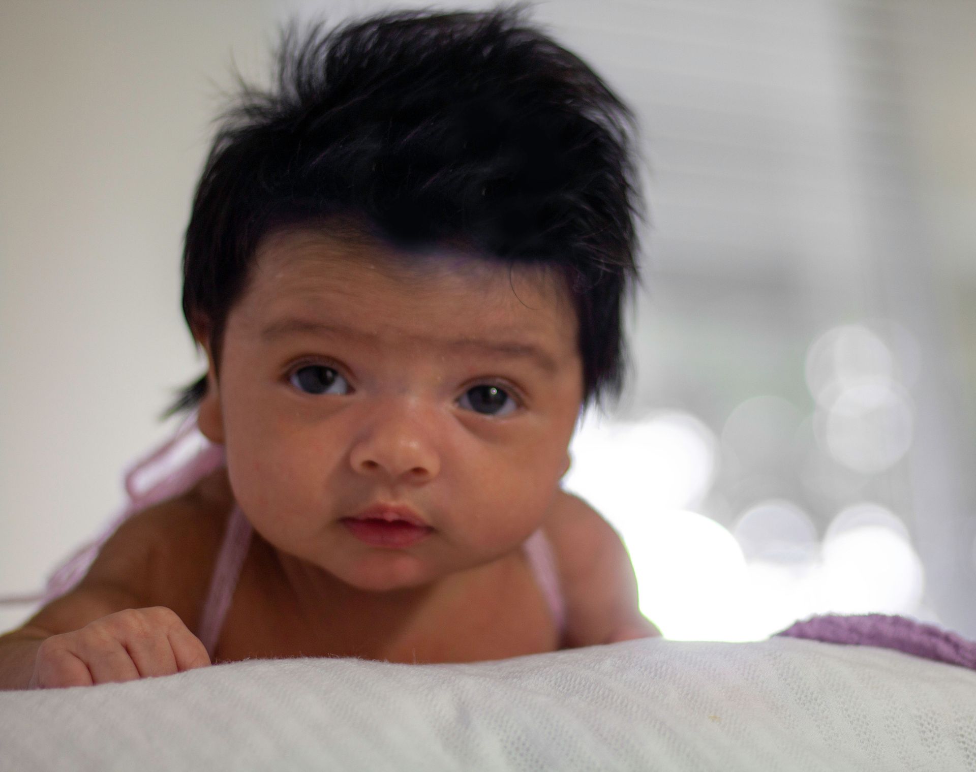 Baby lying on stomach with dark hair, looking intently at the camera.