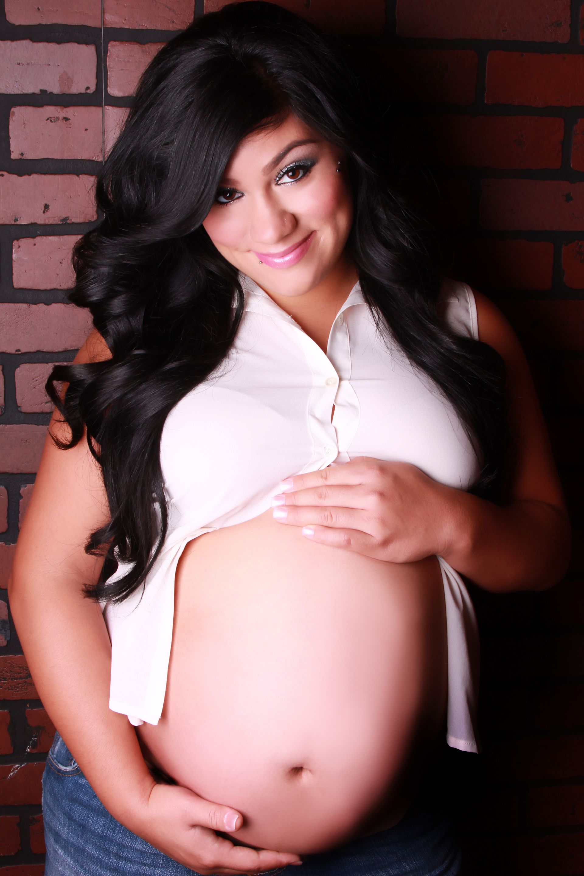 Pregnant woman in a white top and jeans, smiling, hands on her belly, against a brick wall.