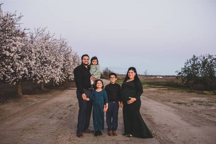 Family of five poses on a dirt path; blossoming trees line the sides. A pregnant woman stands next to her family.