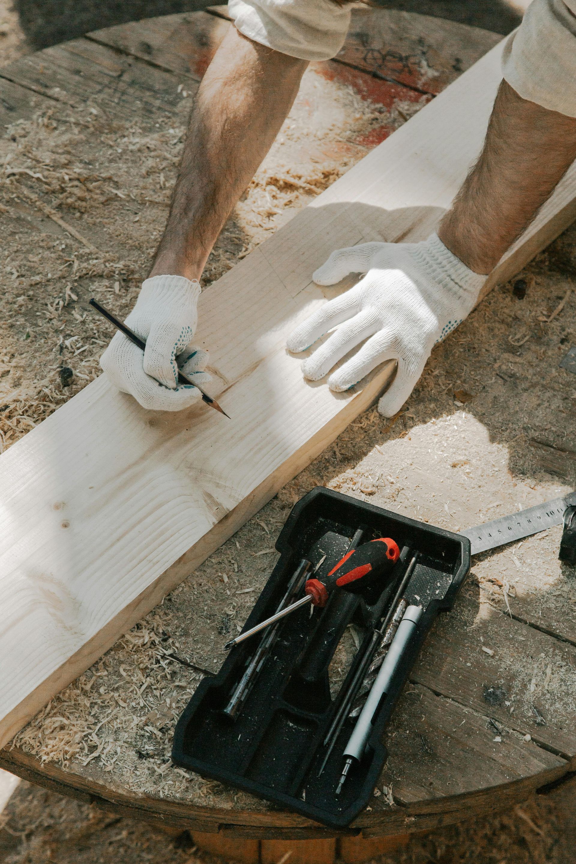 Person in white gloves marking a wooden plank with a pencil beside a toolbox on the floor