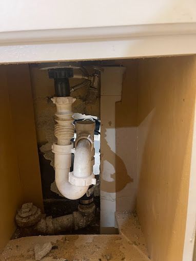 View of plumbing under a sink. White and black pipes are visible. Setting is inside a cabinet with light-colored walls.