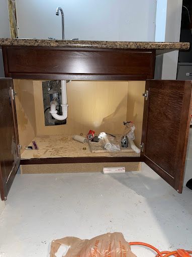 Open cabinet under a sink, with plumbing visible and debris on the floor, in a white-floored room.