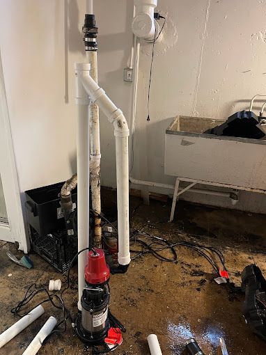 Basement sump pump installation with white PVC pipes, red electrical components, and messy concrete floor.