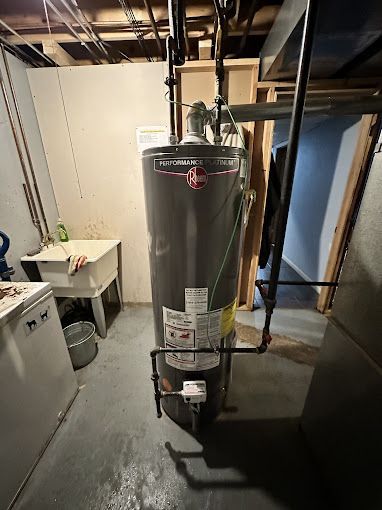 A tall, gray water heater in a basement. Copper pipes and a black pipe extend from it.