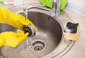 Hands in yellow gloves cleaning kitchen sink drain.