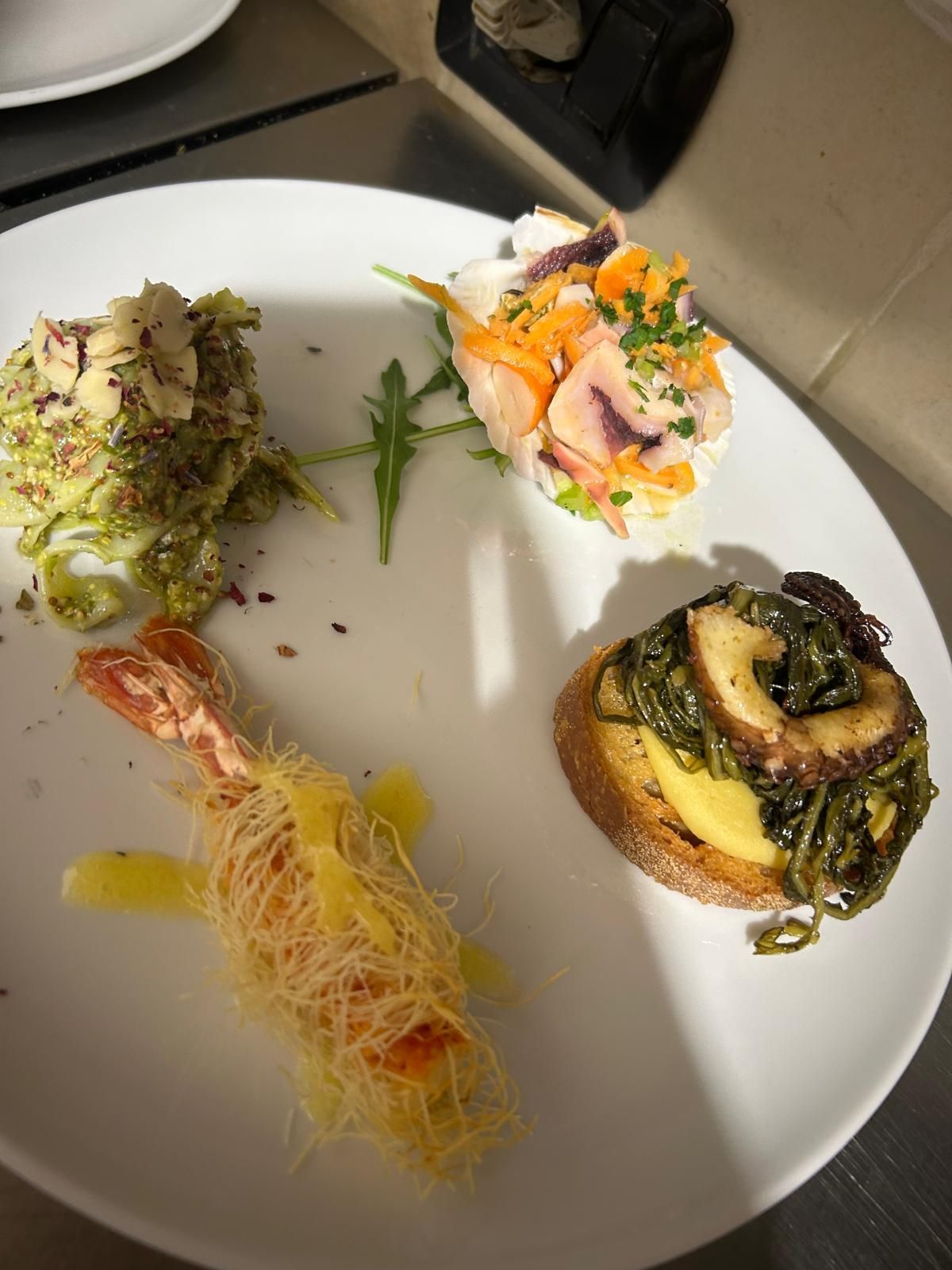 Piatto bianco con quattro piccoli antipasti gourmet, ciascuno guarnito con erbe aromatiche e salsa.