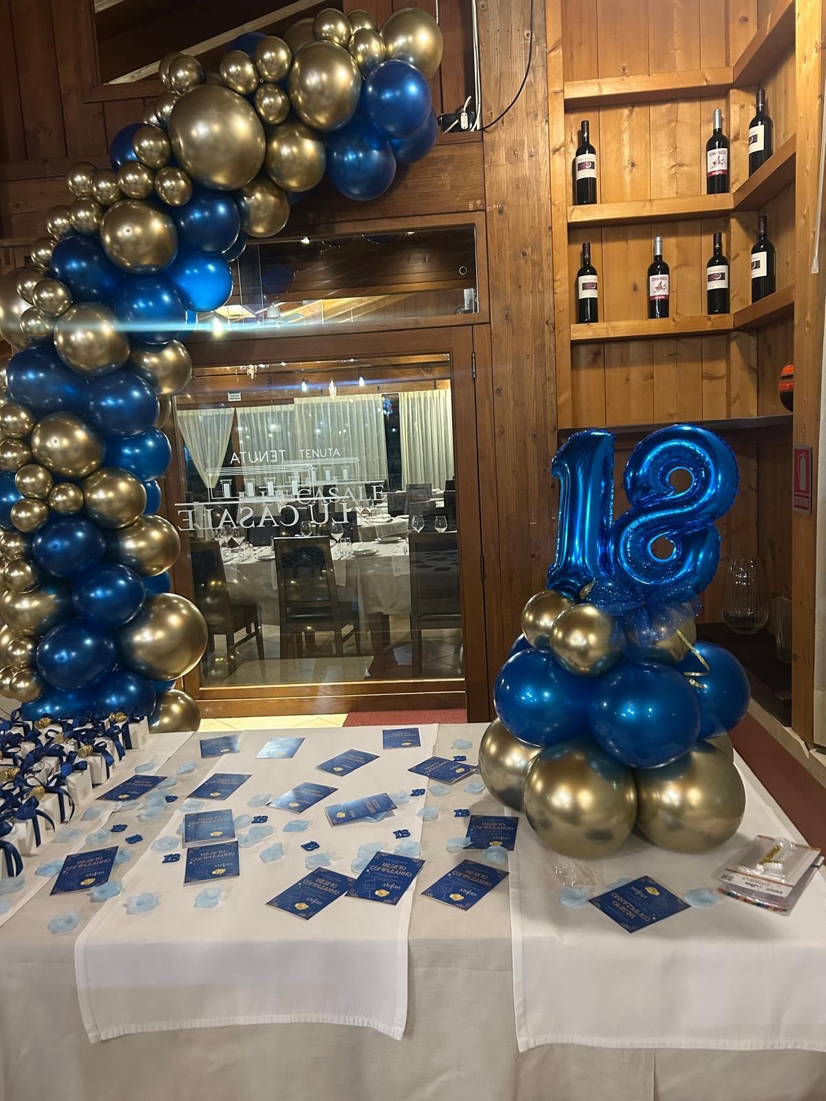 Composizione di palloncini blu e oro sopra un tavolo con volantini all'ingresso di una location rivestita in legno.