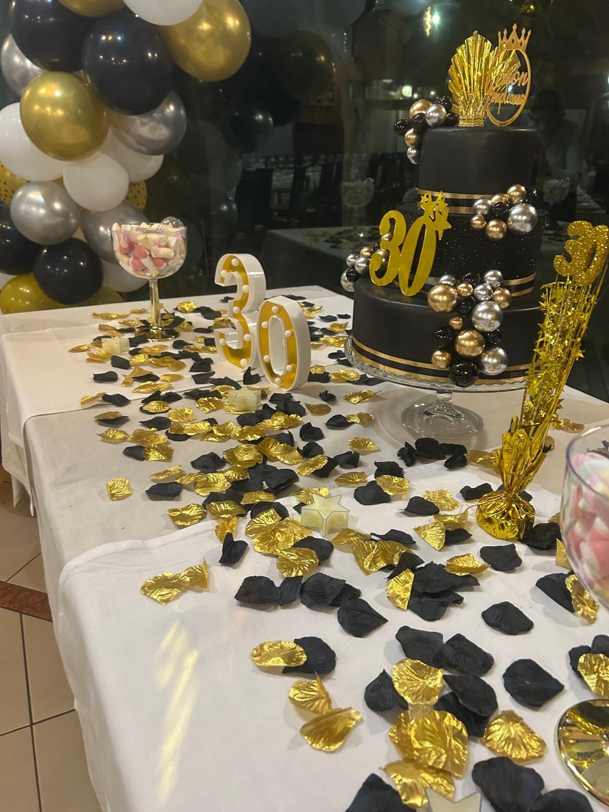 Tavolo per la festa del 30° compleanno in nero e oro con torta, palloncini e coriandoli dorati sparsi.