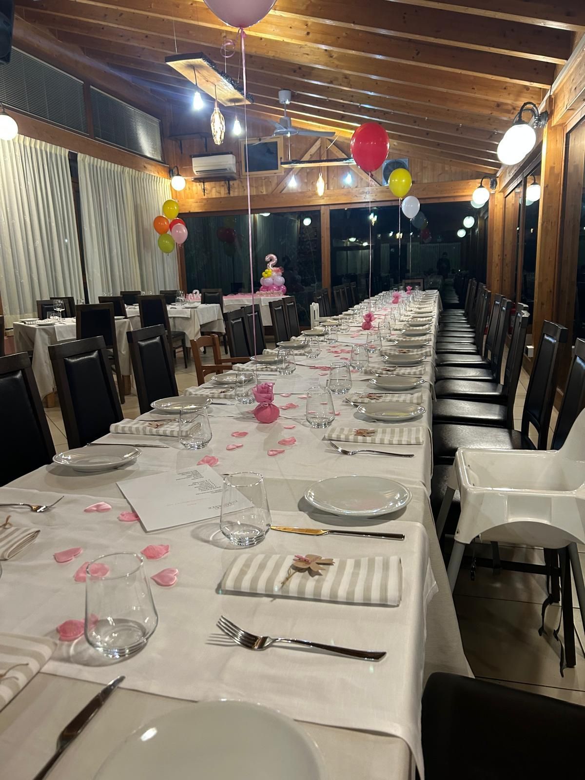 Un lungo tavolo da banchetto allestito per un evento sotto un patio in legno, con palloncini rossi e gialli appesi sopra.