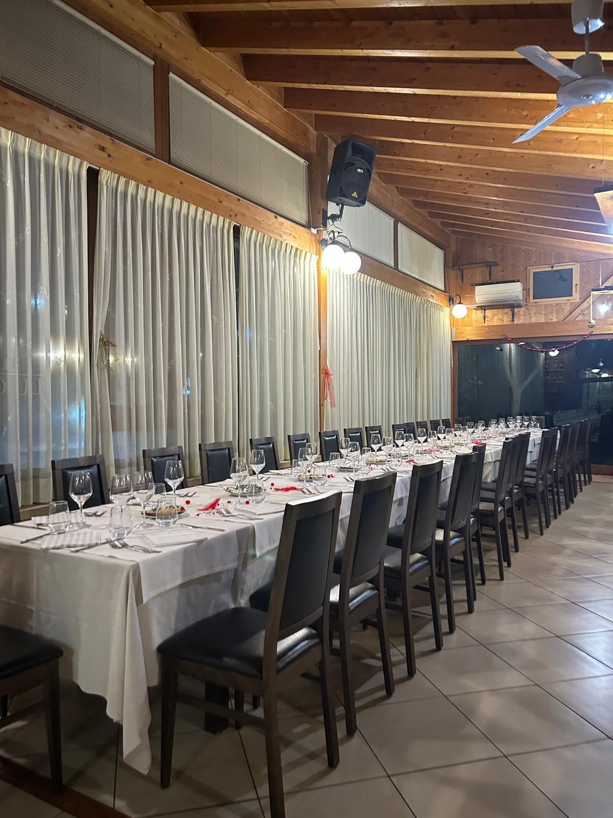 In una sala da pranzo con travi a vista, tende e pavimento piastrellato, è stato allestito un lungo tavolo da banchetto con sedie nere.