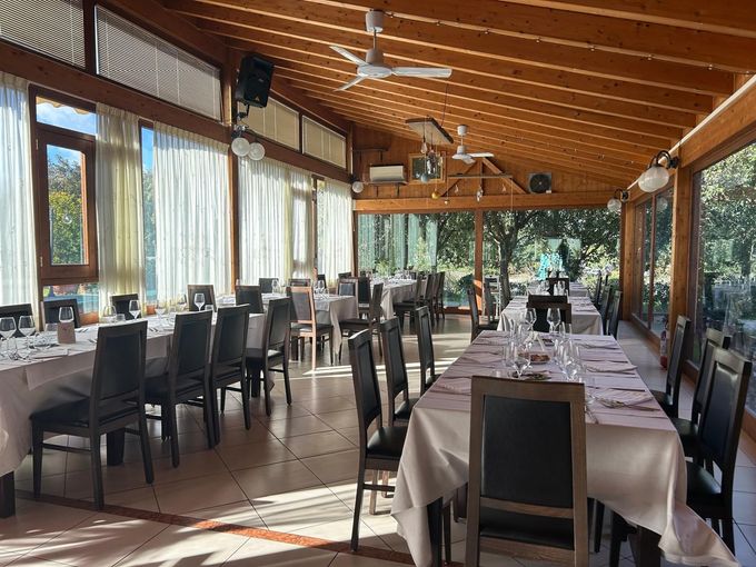 Sala da pranzo vuota di un ristorante con tavoli apparecchiati, sedie scure e grandi finestre sotto un soffitto con travi in legno.