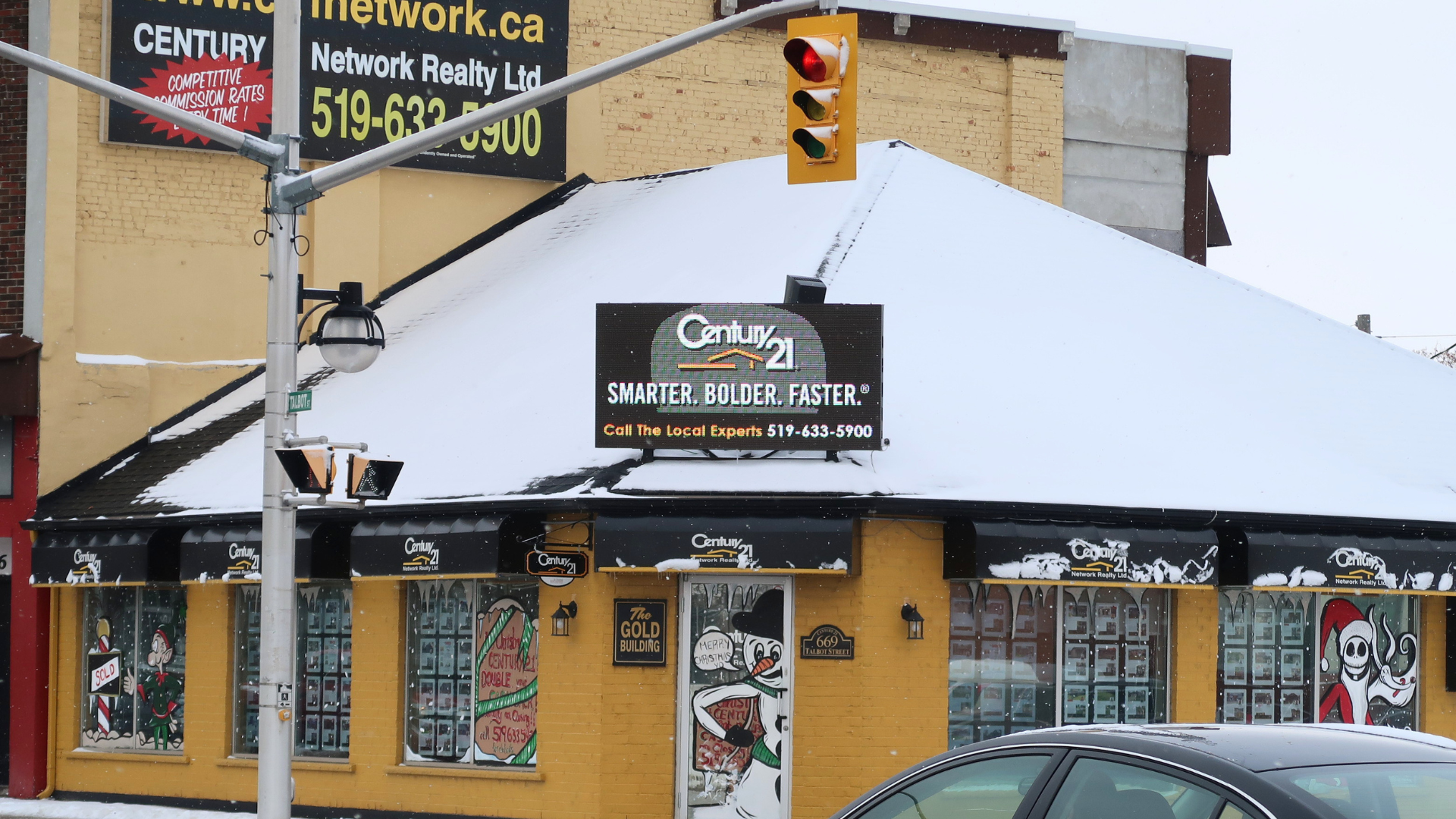 Snow-covered corner storefront with black awning, traffic light, and parked car on a city street.