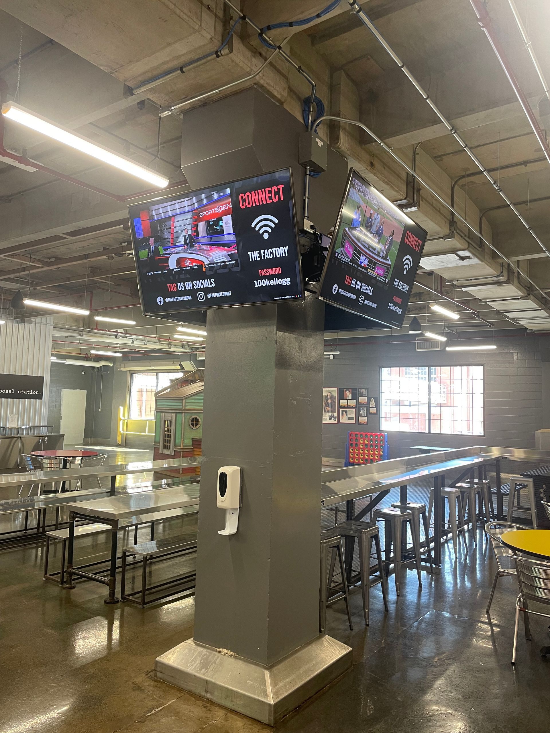 Empty food court with metal tables, central concrete pillar, and overhead TVs under bright industrial lighting