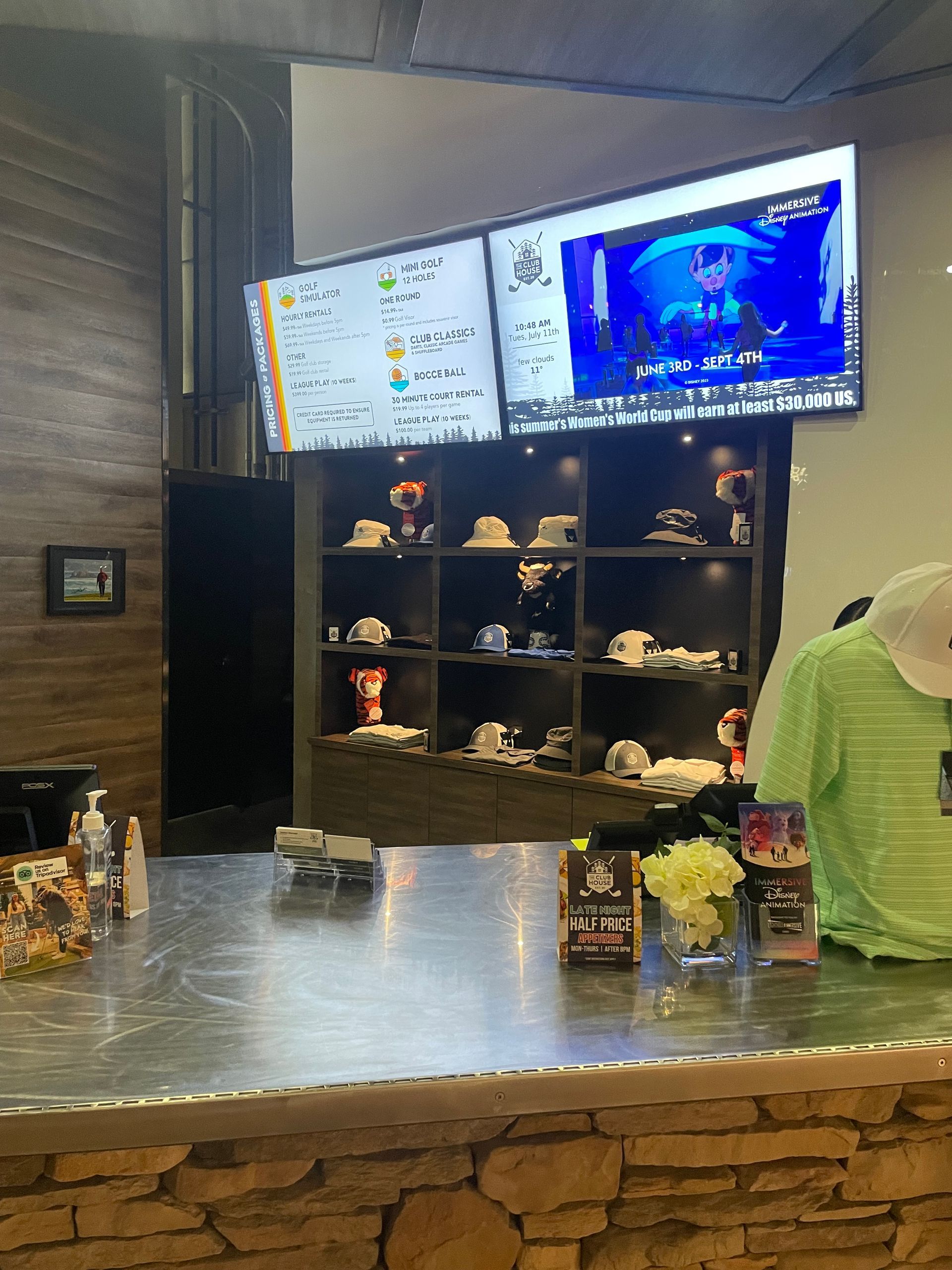 Indoor bar with stone counter, shelves of bottles and decor, and two TV screens overhead