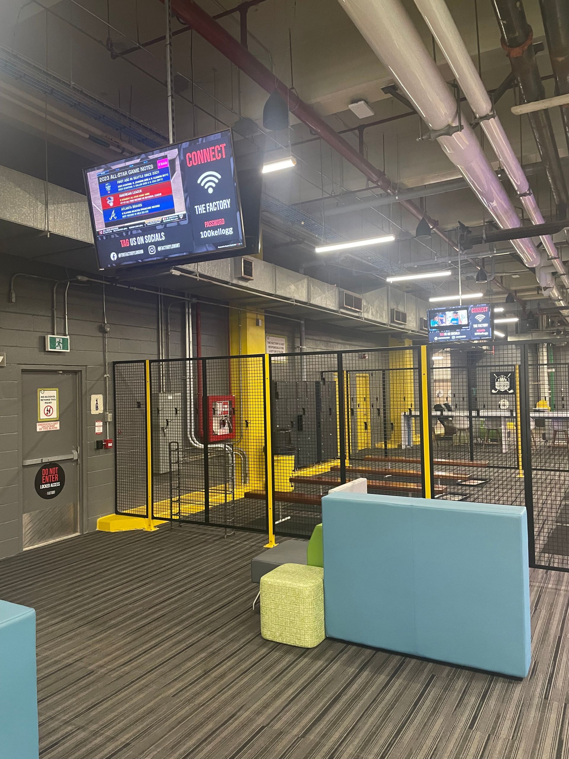 Indoor industrial play area with metal cage walls, yellow trim, carpet, and a blue bench under overhead pipes