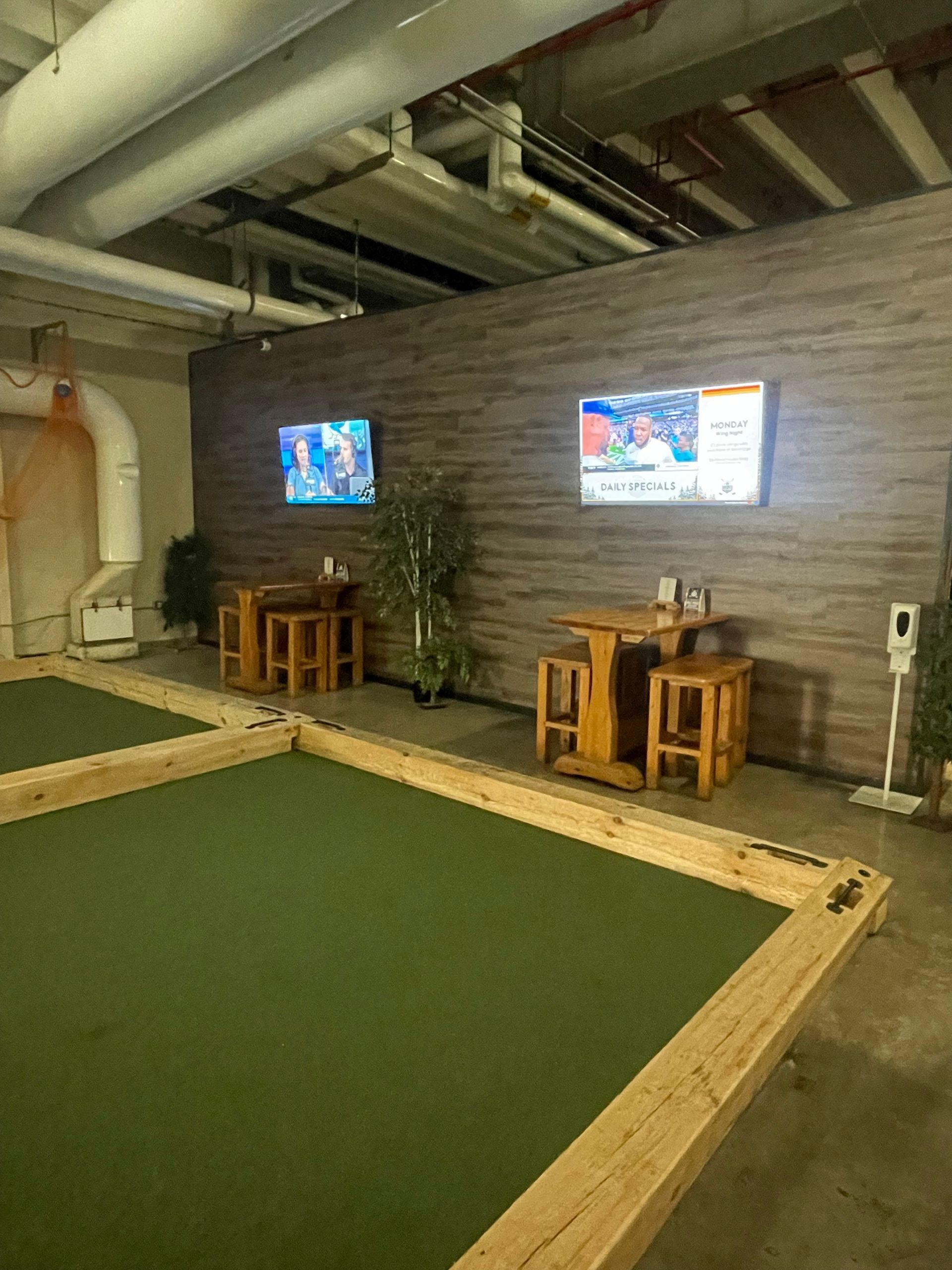 Indoor lounge with green turf floor, two wall-mounted TVs, and wooden stools against a brick wall