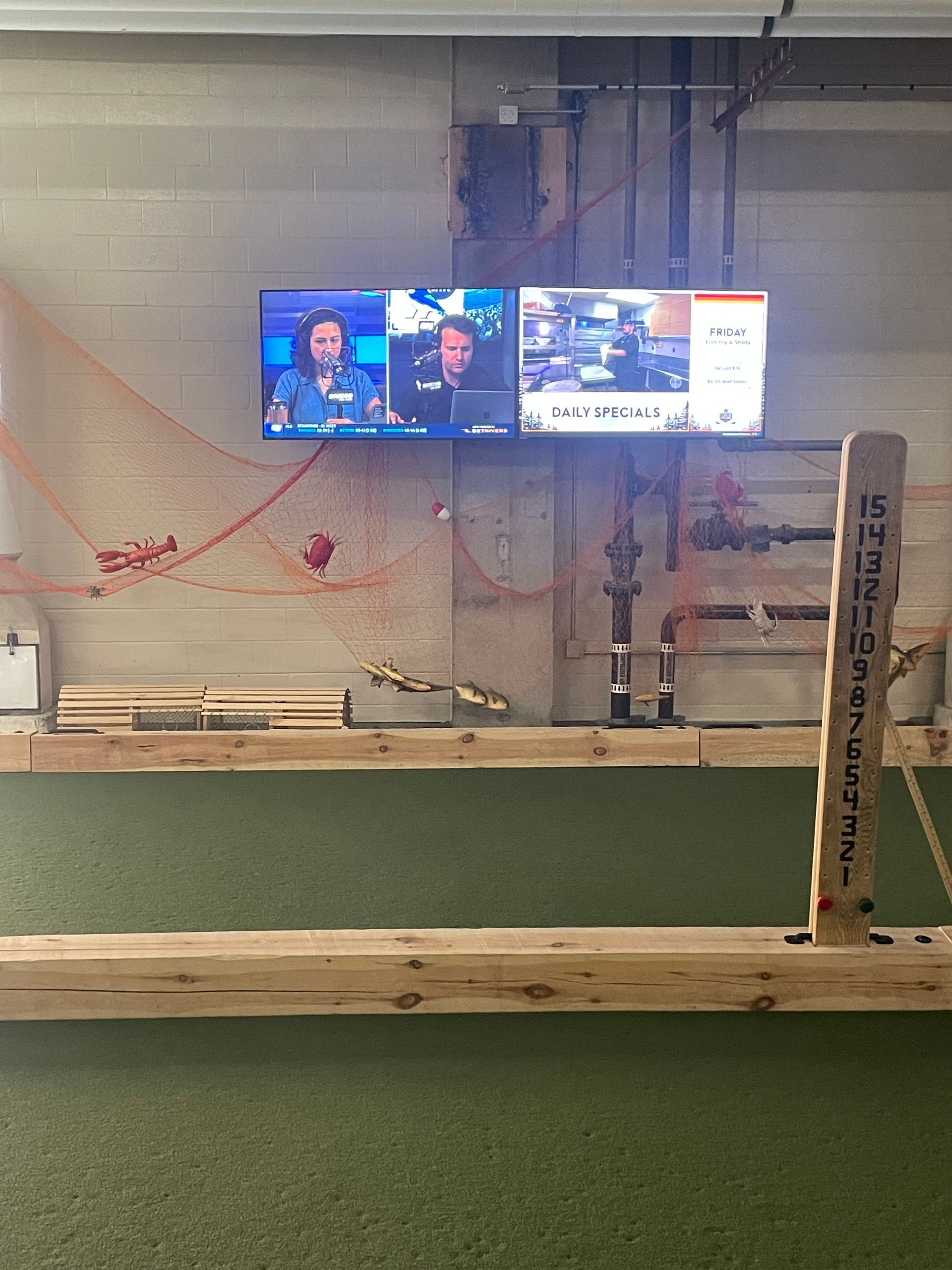 Indoor batting cage with green turf, wooden rail, and wall-mounted TV screens in the background