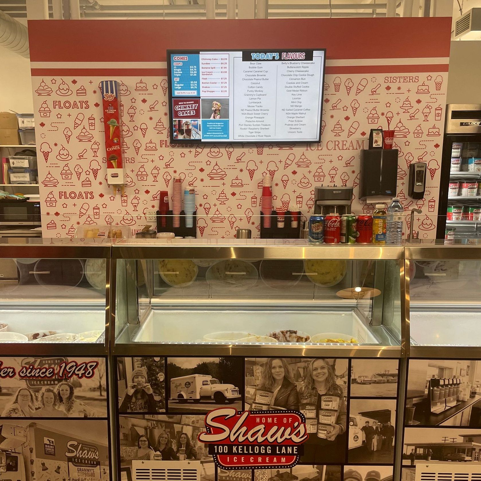 Ice cream counter with menu screens, condiment bottles, and display case inside a Shaw’s Pizza shop.