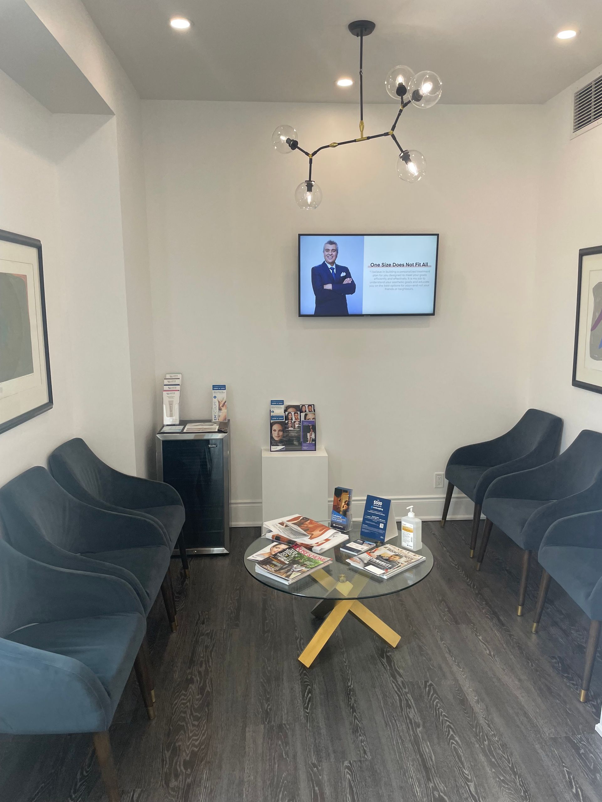 Modern waiting room with dark sofas, a coffee table, wall TV, and chandelier lighting.