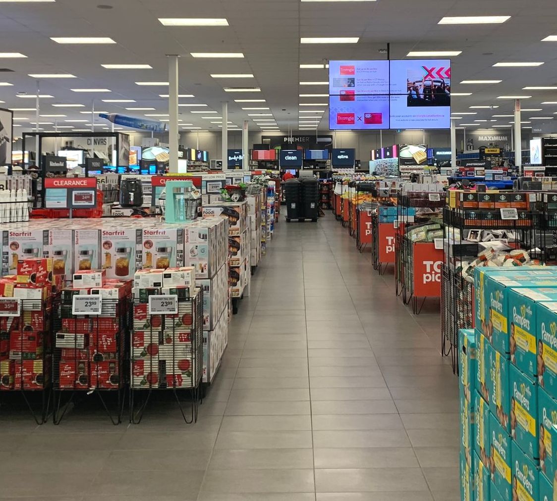 A Canadian Tire store aisle with stacked bulk packages, bright ceiling lights, and digital signs at the back.