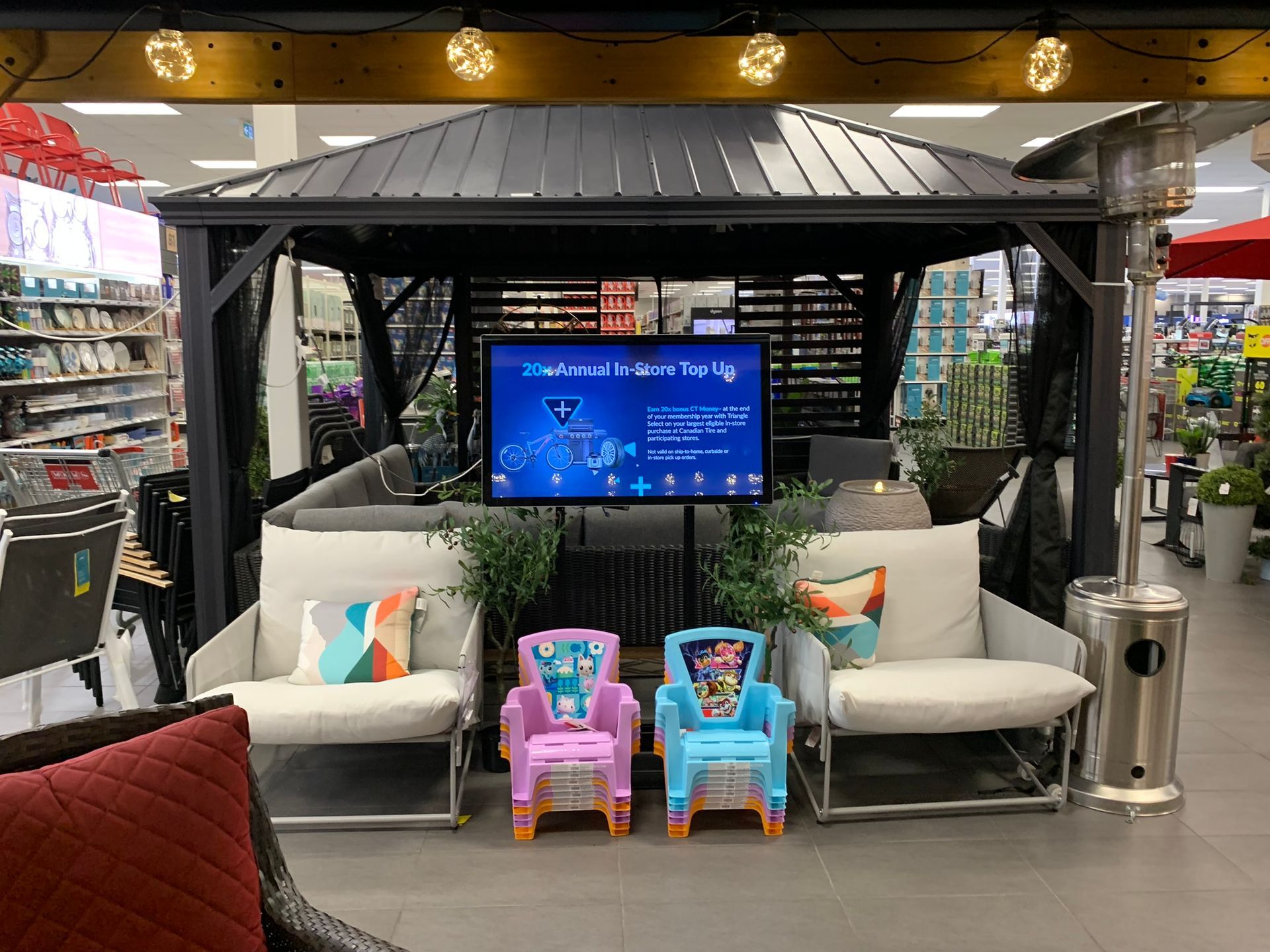Outdoor store display booth with TV, white benches, colorful pillows, and two small toy chairs under a canopy