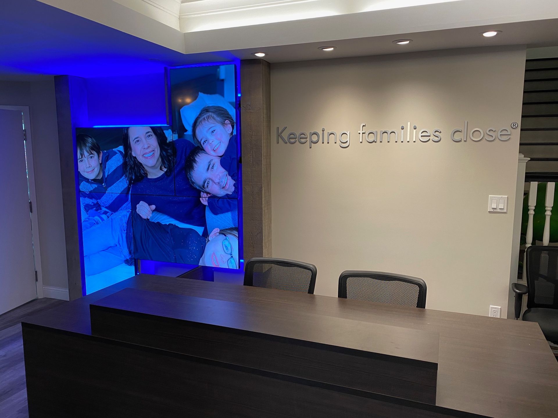 Modern office reception area with blue-lit wall art, desk, and chairs; wall text reads “Keeping families …”