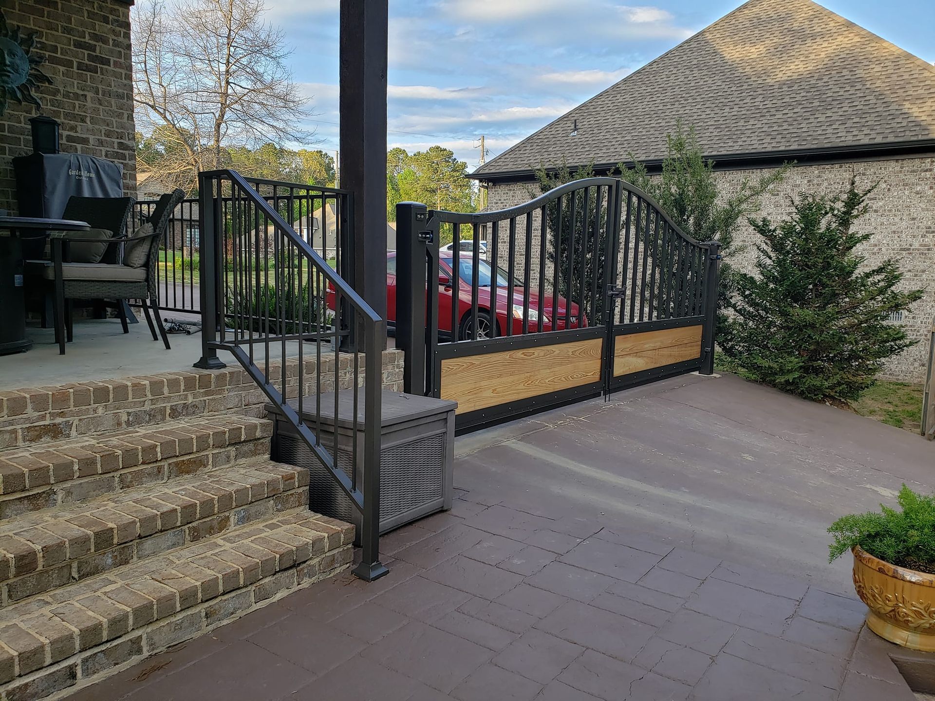 A brick porch with stairs and a metal railing with a red car parked in the background.