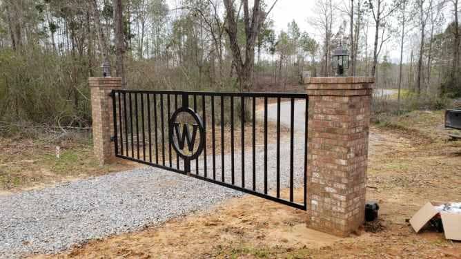 A black gate with the letter w on it is sitting next to a brick wall.