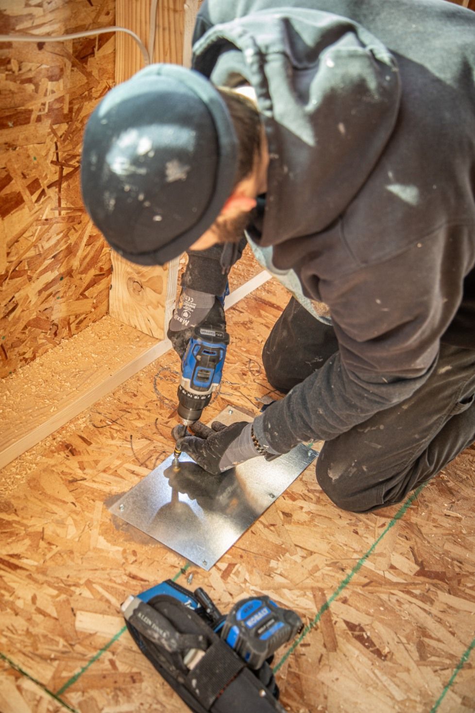 Person kneeling and using a drill to attach a metal sheet to wood framing inside a structure under construction.