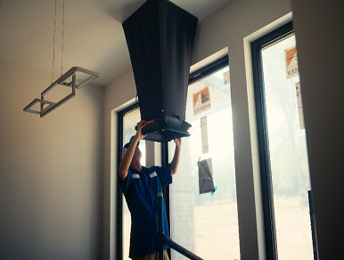 Man installs dark ventilation cover near windows; modern interior.